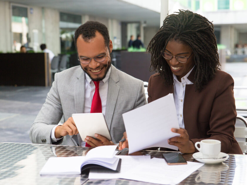 Cheerful business colleagues checking documents during coffee break. Business man and woman sitting in cafe, reading documents and using tablet. Paperwork concept